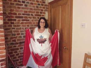 Me- dark hair, pale skin, glasses, smiling- wearing a sleeveless shirt with a maple leaf on it, and I'm holding a Canadian flag. A brick wall and a wood door are behind me.